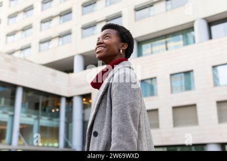 Seitenansicht eines lächelnden jungen Afroamerikaners, der vor verschwommenen Gebäuden im Hintergrund auf der Straße steht Stockfoto