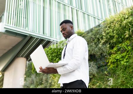 Von unten ein junger afroamerikanischer Freelancer in weißem Hemd, der Netbook hält, während er während der Online-Arbeit in der Nähe von Hecke und Bürogebäuden steht Stockfoto