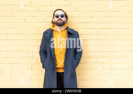 Ein entspannter Mann mit Sonnenbrille und gelbem Hoodie steht selbstbewusst vor einer gelben Ziegelwand und verkörpert urbanen Street Style. Stockfoto