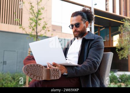 Ein zeitgenössischer Geschäftsmann in modischer Kleidung arbeitet intensiv an seinem Laptop, während er vor einem städtischen Gebäude sitzt. Stockfoto