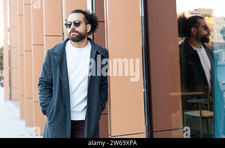 Ein modischer Mann mit Sonnenbrille und Bart lehnt sich an die Wand eines modernen Gebäudes und reflektiert auf dem Glas. Stockfoto