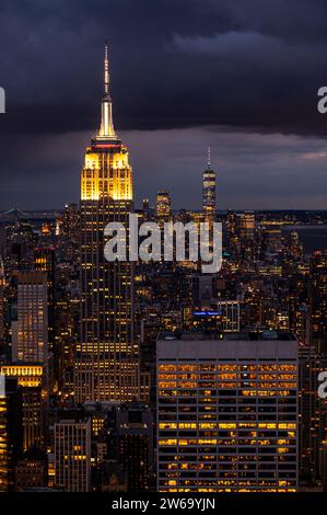 Blick aus der Vogelperspektive auf die Stadt mit beleuchteten Wolkenkratzern und das Empire State Building mit Hochhäusern vor dem bewölkten Abendhimmel in New York City Stockfoto