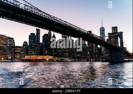 Spektakuläre Stadtlandschaft von Manhattan mit Wolkenkratzern und Brooklyn Bridge über dem ruhigen Fluss bei Nacht in New York City unter blauem wolkenlosem Himmel Stockfoto