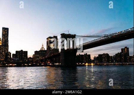 Spektakuläre Stadtlandschaft von Manhattan mit Wolkenkratzern und Brooklyn Bridge über dem ruhigen Fluss in der Dämmerung in New York City unter blauer Wolke Stockfoto