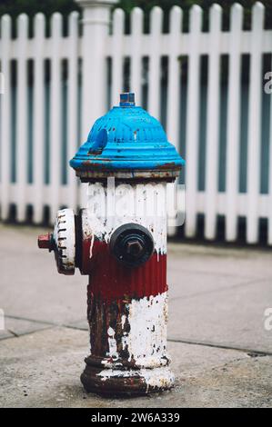 Ein klassischer blauer und roter Hydrant steht vor einem weißen Streifenzaun in Manhattan, New York. Titel: Klassischer Feuerhydrant in man Stockfoto