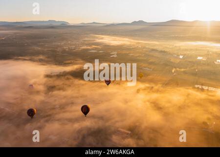 Von oben erheben sich mehrere bunte Heißluftballons über einer nebelverhangenen Landschaft, während die Sonne ein sanftes Leuchten auf die Szene wirft Stockfoto