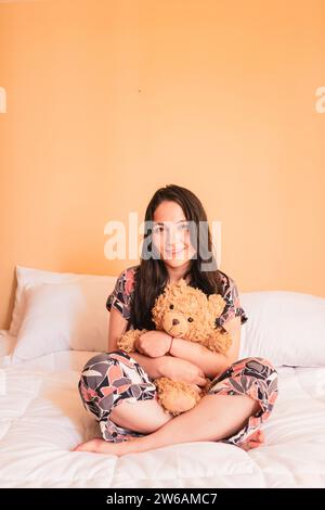 Voller Körper einer glücklichen jungen Frau im Pyjama und langen Haaren, die in die Kamera schaut, während sie mit überkreuzten Beinen auf dem Bett sitzt und sich in weichem, weichem, braunem Teddybär umgibt Stockfoto