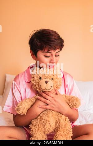 Glückliche junge Frau im Pyjama und kurzen Haaren mit geschlossenen Augen, während sie auf dem Bett sitzt und sich an den weichen, weichen, braunen Teddybären anschmiegt Stockfoto