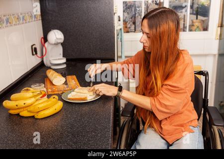 Eine unabhängige Frau im Rollstuhl sieht man, wie sie in einer gut ausgestatteten, gemütlichen Küche Brot für ein Sandwich schneidet. Stockfoto