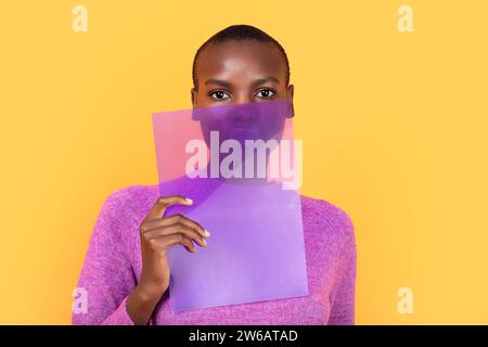 Porträt einer afroamerikanischen jungen Frau in rosa Pullover mit transparentem Plastik, während sie auf die Kamera vor gelbem Hintergrund blickt Stockfoto