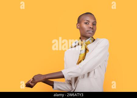 Selbstbewusste afroamerikanische schlanke junge Frau in modischer Freizeitkleidung, die einen Schal um den Hals trägt, während sie die Hände vor dem gelben Hintergras hält Stockfoto