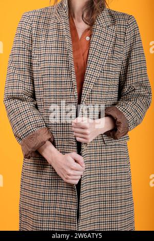 Abgeschnittene, nicht erkennbare weiße schlanke Frau mit kariertem Muster, langer Mantel, der in die Kamera blickt, während sie vor gelbem Hintergrund steht Stockfoto