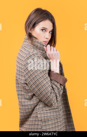 Seitenansicht einer attraktiven jungen kaukasischen schlanken Frau mit kariertem Muster, die in die Kamera blickt, während sie vor gelbem Hintergrund steht Stockfoto