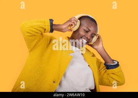 Fröhliche afroamerikanische junge Frau, die in der Jacke nach unten blickt und Musik über kabellose Kopfhörer vor gelbem Hintergrund hört Stockfoto