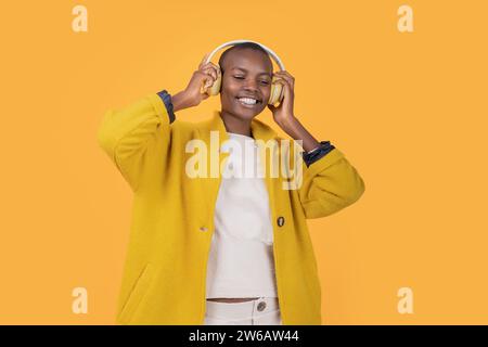 Fröhliche afroamerikanische junge Frau, die in der Jacke nach unten blickt und Musik über kabellose Kopfhörer vor gelbem Hintergrund hört Stockfoto