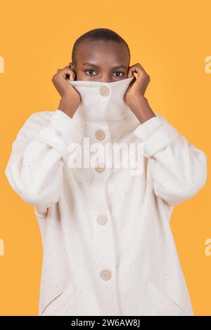Afroamerikanische junge Frau in trendiger Rollkragenbekleidung, die den Mund bedeckt und die Kamera vor gelbem Hintergrund betrachtet Stockfoto