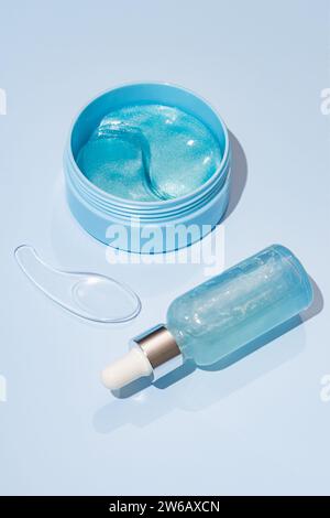 Eine Draufsicht eines blauen Gels in einem offenen Glas neben einer Flasche mit Tropfer, die beide Hautpflegeprodukte auf pastellblauem Hintergrund enthalten. Stockfoto