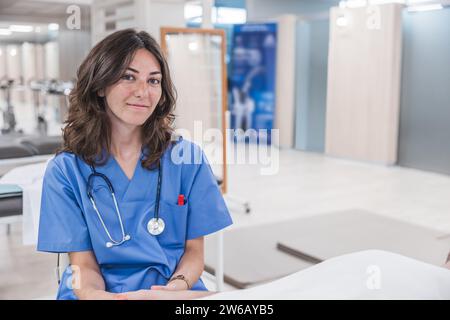 Lächelnde junge Ärztin mit Stethoskop um den Hals und in Uniform, die in die Kamera blickt, während sie am Tisch im Krankenhaus vor verschwommenem Innenraum sitzt Stockfoto