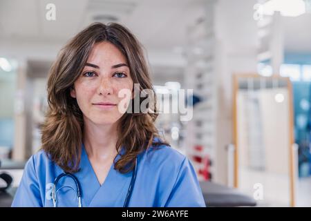 Portrait junge Ärztin mit Stethoskop um den Hals und in Uniform in die Kamera beim Sitzen am Tisch im Krankenhaus gegen verschwommenes Inneres Stockfoto