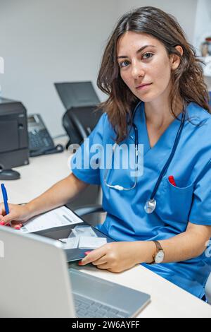 Glückliche junge Ärztin in Uniform und mit Stethoskop, die in die Kamera schaut, während sie im Sprechzimmer am Tisch sitzt und mit Laptop das Handy hält Stockfoto