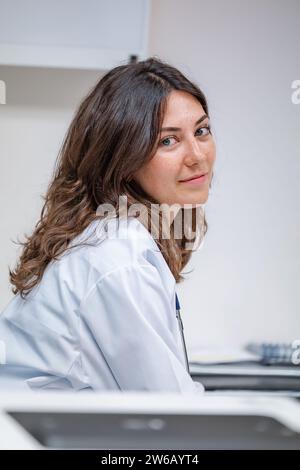 Porträt einer jungen Ärztin in medizinischer Uniform und Stethoskop, die im Krankenhaus vor weißem Hintergrund sitzt, während sie in die Kamera schaut Stockfoto