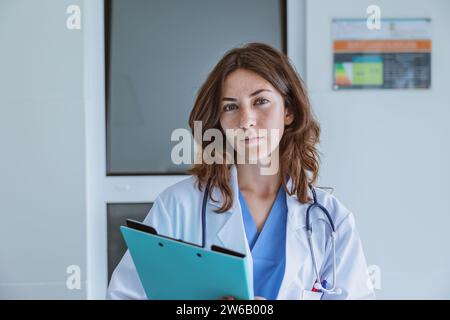 Porträt einer jungen Ärztin in Uniform und Stethoskop, die mit Mappe im Krankenhaus steht, während sie die Kamera gegen die weiße Wand sieht Stockfoto