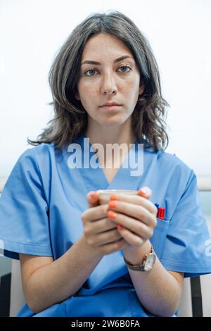 Junge Ärztin in blauer Uniform, die in die Kamera schaut, während sie die Tasse in den Händen hält und auf dem Stuhl vor weißem Hintergrund sitzt Stockfoto