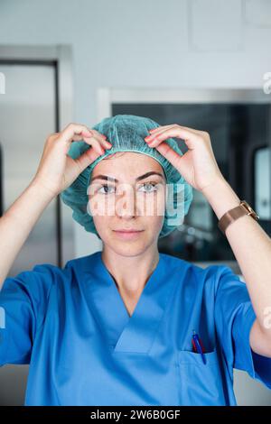 Junge Chirurgin in blauer Uniform und Kappe, die in die Kamera blickt, während sie vor verschwommenem Hintergrund des Korridors steht Stockfoto