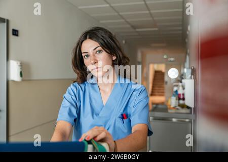 Junge Ärztin mit blauer Uniform und Blick in die Kamera, während sie im modernen Klinikkorridor vor verschwommenem Hintergrund sitzt Stockfoto