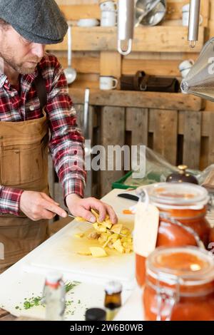 Der auf die Ernte fokussierte Koch in Schürze und Baskenmütze steht in einer rustikalen Küche und schneidet aufmerksam Kartoffeln auf einem weißen Schneidebrett mit einem Hintergrund aus hängendem Nuss Stockfoto