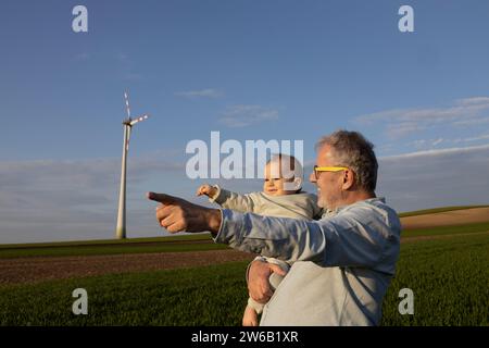 Glücklicher Großvater, der dem süßen Jungen etwas zeigt, während er auf der Windfarm vor blauem Himmel steht Stockfoto