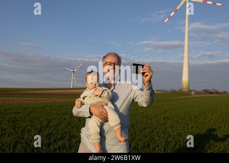 Glücklicher Großvater macht Selfie mit Enkel über Smartphone auf Windpark gegen Himmel Stockfoto