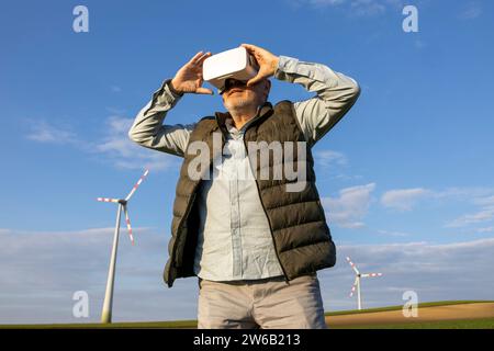 Niedriger Blickwinkel eines älteren Mannes, der eine VR-Brille trägt, während er mit erhobenen, ausgestreckten Armen gegen die Windturbine und den blauen Himmel steht Stockfoto