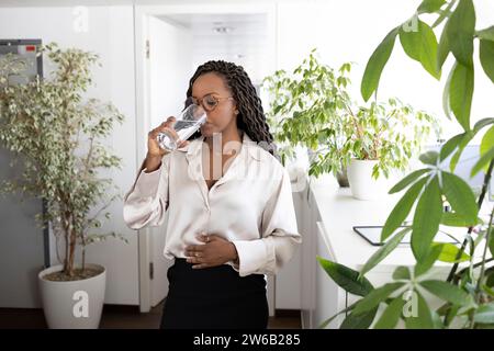 Junge afroamerikanische Geschäftsfrau trinkt Wasser aus Glas, während sie im Büro mit der Hand auf dem Bauch steht Stockfoto