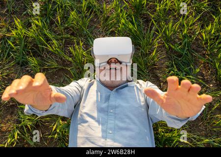 Direkt über dem Schuss eines fröhlichen älteren Mannes mit VR-Brille, während er an sonnigem Tag auf dem Gras auf der Farm liegt Stockfoto