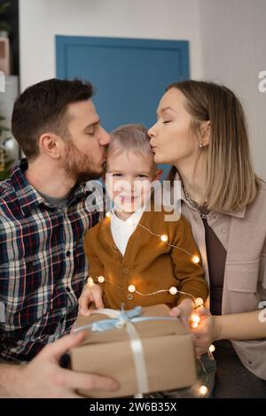 Ein junges Paar mit geschlossenen Augen, das zu Hause vor einem festlichen Weihnachtsbaum sitzt, gibt ihrem Kind liebevoll einen Kuss auf die Wange, während er guckt Stockfoto