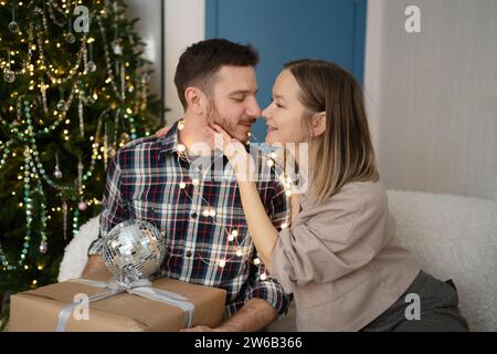 Lächelndes junges Paar, das auf dem Sofa sitzt und Geschenke und liebevolle Gesten vor einem wunderschön geschmückten Weihnachtsbaum zu Hause austauscht Stockfoto