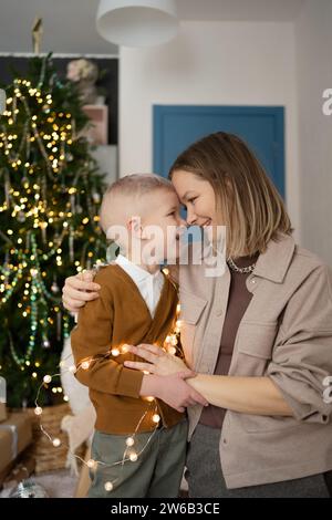 Zärtlicher Moment zwischen einer Mutter und ihrem Sohn, ihre Köpfe berühren sich liebevoll, während sie in Weihnachtslichter gehüllt sind, mit der Wärme Christi Stockfoto