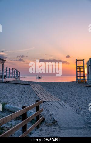Leerer Holzsteg am Sandstrand vor dem Segelboot, das während des Sonnenuntergangs auf Sardinien, Italien, auf dem ruhigen Meer unter dem Himmel schwimmt Stockfoto