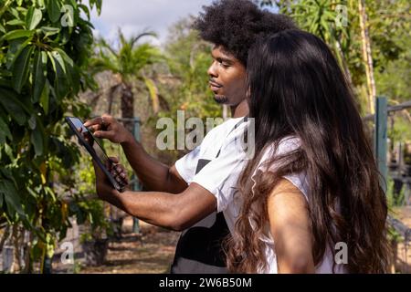 Seitenansicht eines selbstbewussten afroamerikanischen Botanikers, der ein digitales Tablet teilt und mit einer hispanischen Kollegin im Bio-Gewächshaus diskutiert Stockfoto