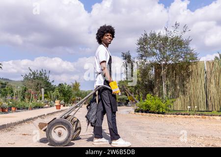 Seitenansicht in voller Länge eines lächelnden jungen afroamerikanischen männlichen Botanikers in Schürze, der mit dem Wagen vor dem Gewächshaus vor dem bewölkten Himmel steht und sich anschaut Stockfoto