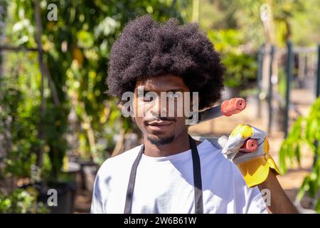 Porträt eines selbstbewusst lächelnden jungen afroamerikanischen Agrarwissenschaftlers mit schwarzem Lockenhaar in schwarzer Schürze, der im Bio-Gewächshaus Haarschneider hält Stockfoto