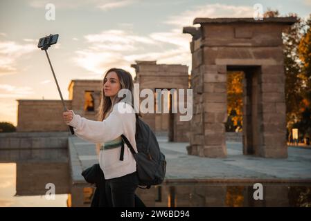 Eine junge Frau nimmt ein Selfie mit ihrem Handy auf einem Selfie-Stick auf, mit der Kulisse des Debod-Tempels in Madrid während des Sonnenuntergangs Stockfoto