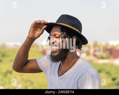 Lächelnder junger bärtiger Afroamerikaner in weißem T-Shirt, der einen schwarzen Hut anpasst und glücklich über das Stadtbild lacht Stockfoto