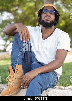 Junge coole bärtige Afroamerikaner in Street-Style-Outfit und Sonnenbrille, die auf Steinrand vor Bäumen sitzt Stockfoto