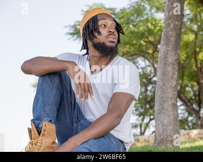 Junge coole bärtige Afroamerikaner im Straßenstil, der auf Steinrand sitzt und wegblickt Stockfoto