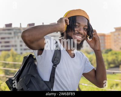 Lächelnder junger afroamerikanischer männlicher Hipster mit Rucksack, der am Sommertag im Stadtpark steht und Kopfhörer berührt und Musik hört Stockfoto