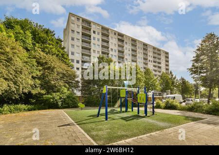 Schaukeln und rutschen Sie auf dem Spielplatz mit Bäumen und Pflanzen vor dem großen Gebäude vor blauem Himmel an sonnigen Tagen Stockfoto