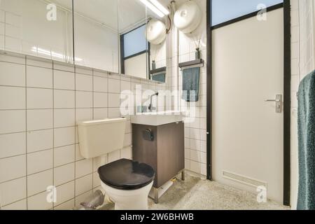 Ein gut beleuchtetes, modernes, kompaktes Badezimmer mit sauberen weißen Fliesen, einem Kosmetikbereich mit schwarzem Wasserhahn und einer Toilette. Stockfoto