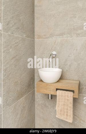 Wasserhahn an der Wand über weißem Keramikwaschbecken auf Holzregal mit Handtuch im Badezimmer Stockfoto